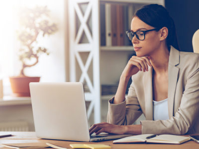 Pensive young beautiful businesswoman in glasses working on laptop and keeping hand on chin while sitting at her working place